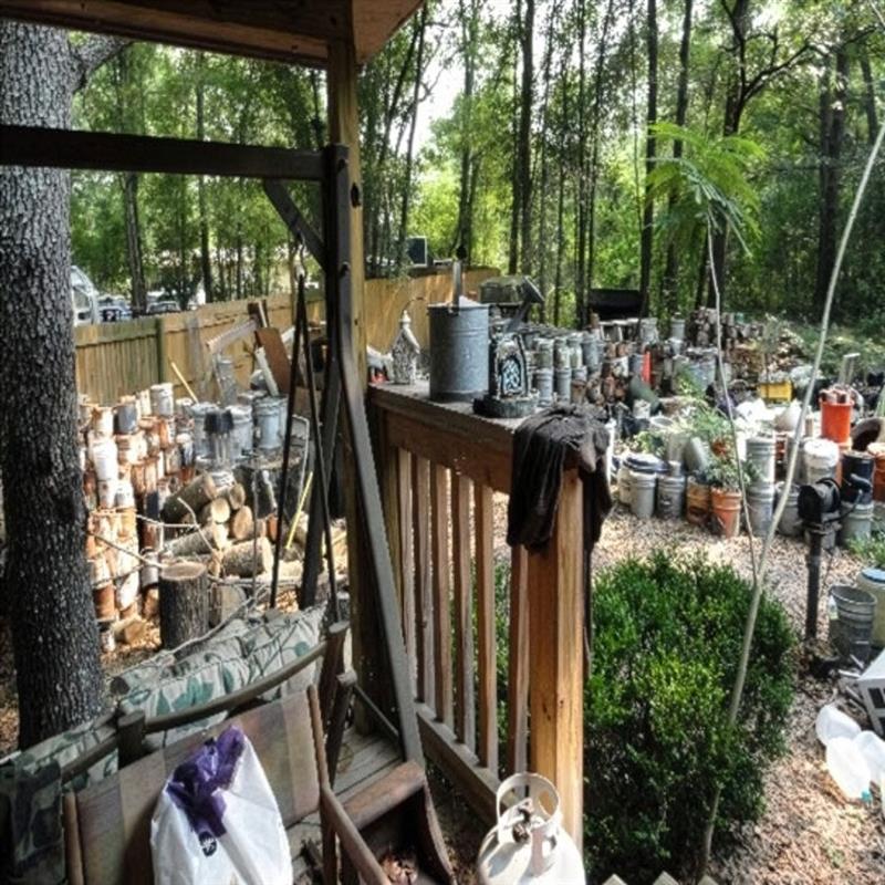 Cluttered backyard beside a wooden porch filled with many metal containers, buckets, and stacked firewood among trees.